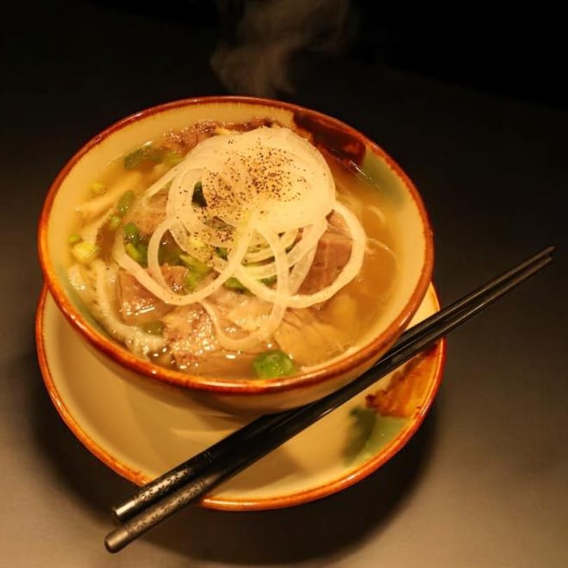 Steaming bowl of Vietnamese pho with beef, noodles, onions, and green herbs, with chopsticks.