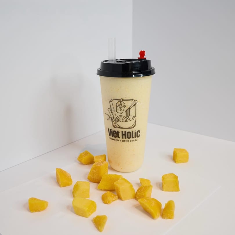 Viet Holic mango smoothie with a straw and red heart stopper, surrounded by mango chunks.